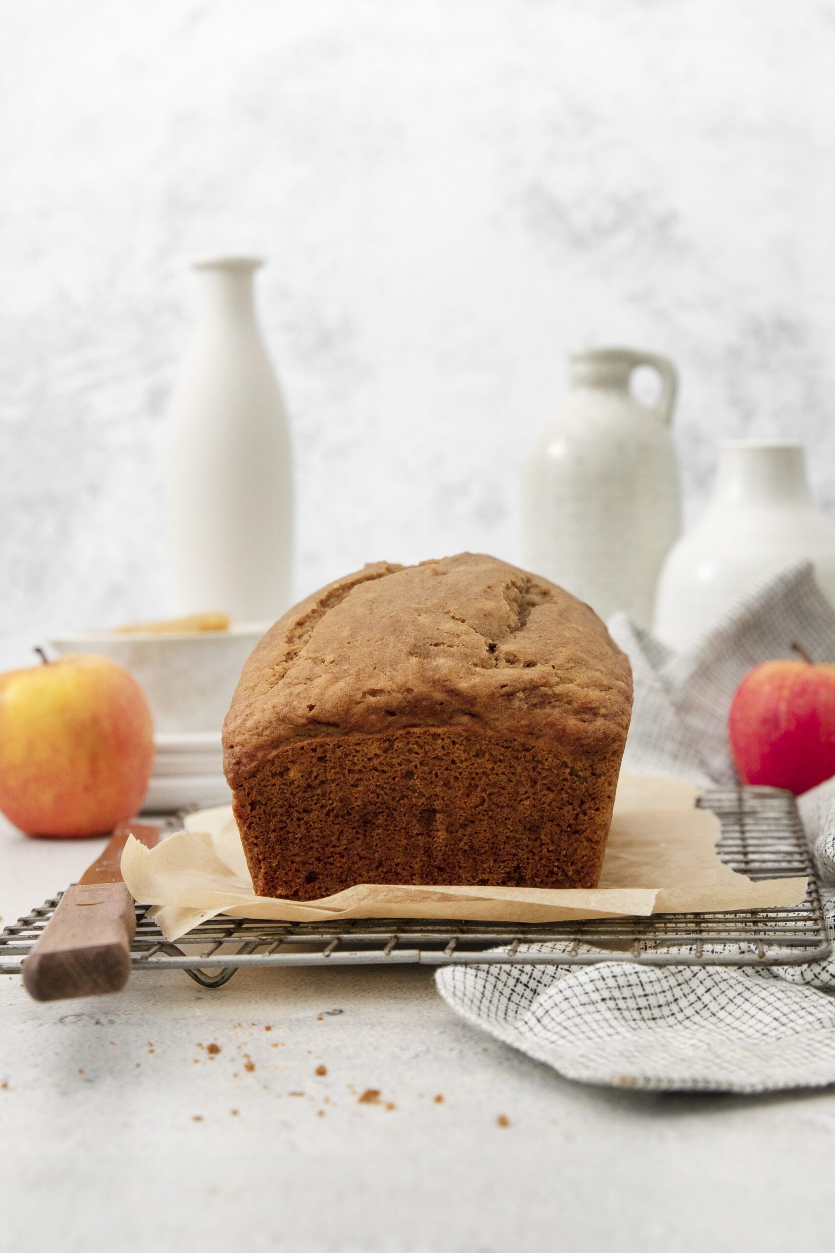 A loaf of apple bread sits on parchment paper on a cooling rack. Two apples, a knife, and ceramic jars are arranged in the background on a light-colored surface. A cloth napkin lies nearby.