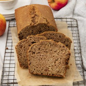 A loaf of apple bread with three slices cut, resting on parchment paper atop a cooling rack. Apples, a cup of applesauce, and a cloth are in the background on a light surface.