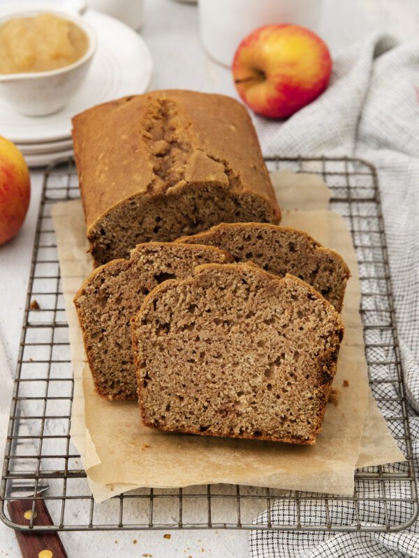 A loaf of apple bread with three slices cut, resting on parchment paper atop a cooling rack. Apples, a cup of applesauce, and a cloth are in the background on a light surface.