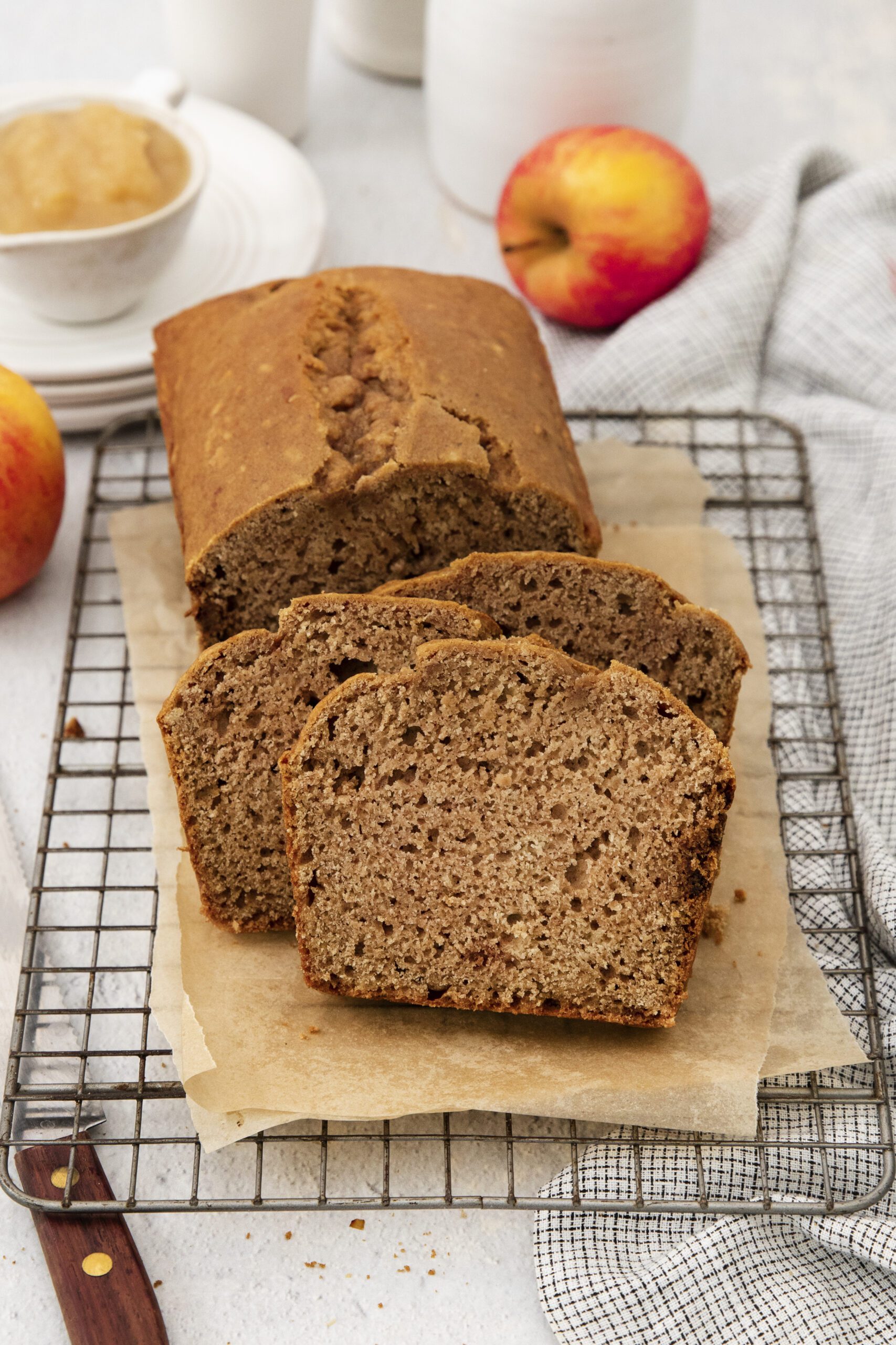 A loaf of apple bread with three slices cut sits on parchment paper atop a cooling rack. Nearby are apples, a cup of applesauce, a knife, and a gray cloth. The background is light and airy.