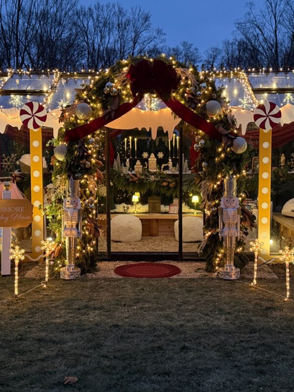 A glass house decorated for Christmas with lights, garlands, large red bows, candy canes, and nutcracker figures. The inside glows warmly with cozy seating and festive decorations. Trees and snowflakes light up the yard.
