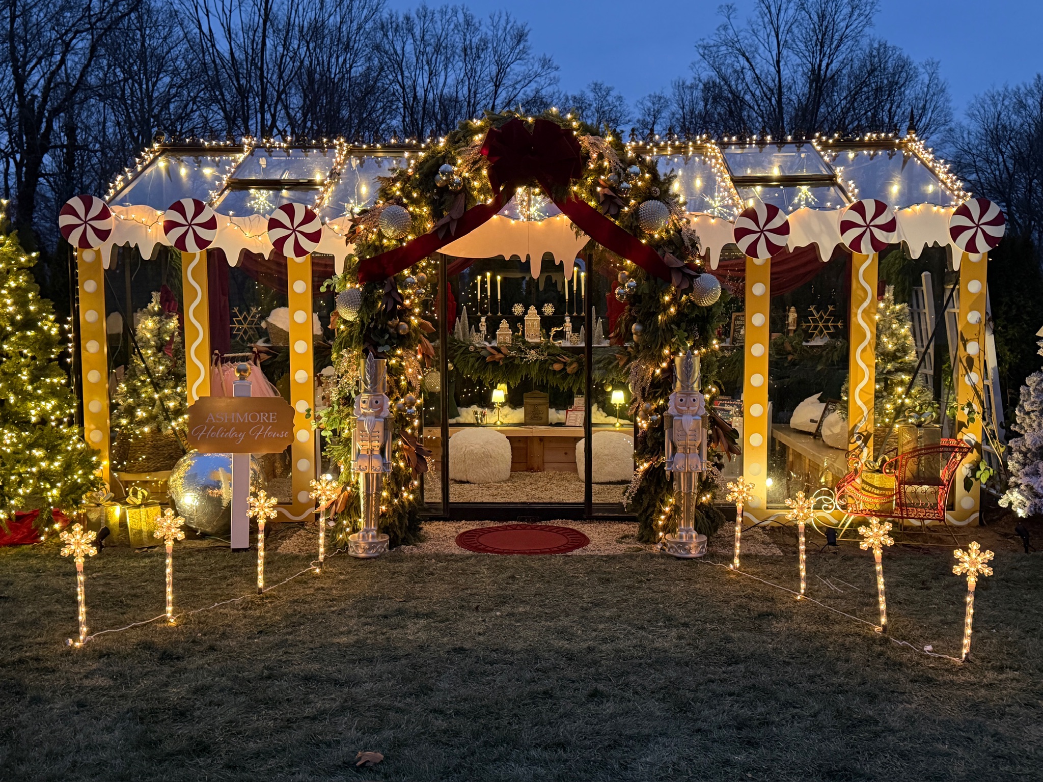 A glass house decorated for Christmas with lights, garlands, large red bows, candy canes, and nutcracker figures. The inside glows warmly with cozy seating and festive decorations. Trees and snowflakes light up the yard.