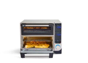 A countertop convection oven with a digital display shows roasted potato wedges on a tray in the open lower compartment and cooked food in the upper compartment, against a plain white background.