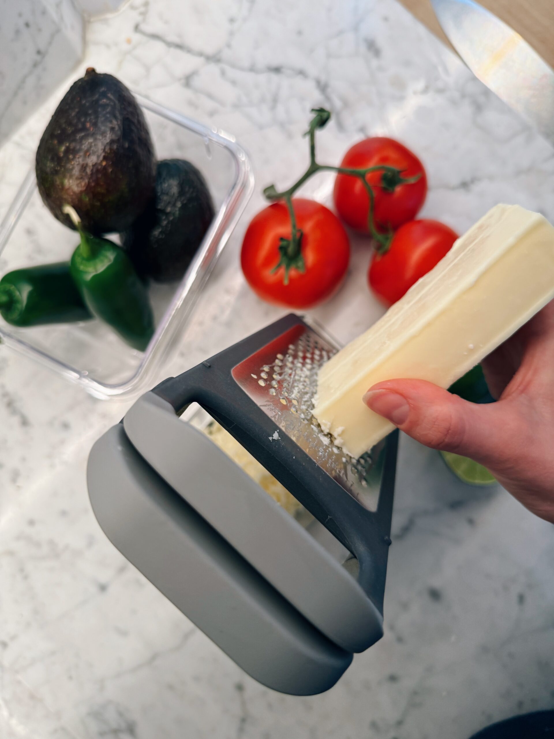 A hand grates a block of cheese over a gray grater on a marble countertop. Nearby are avocados, a green chili pepper in a container, and vine tomatoes.