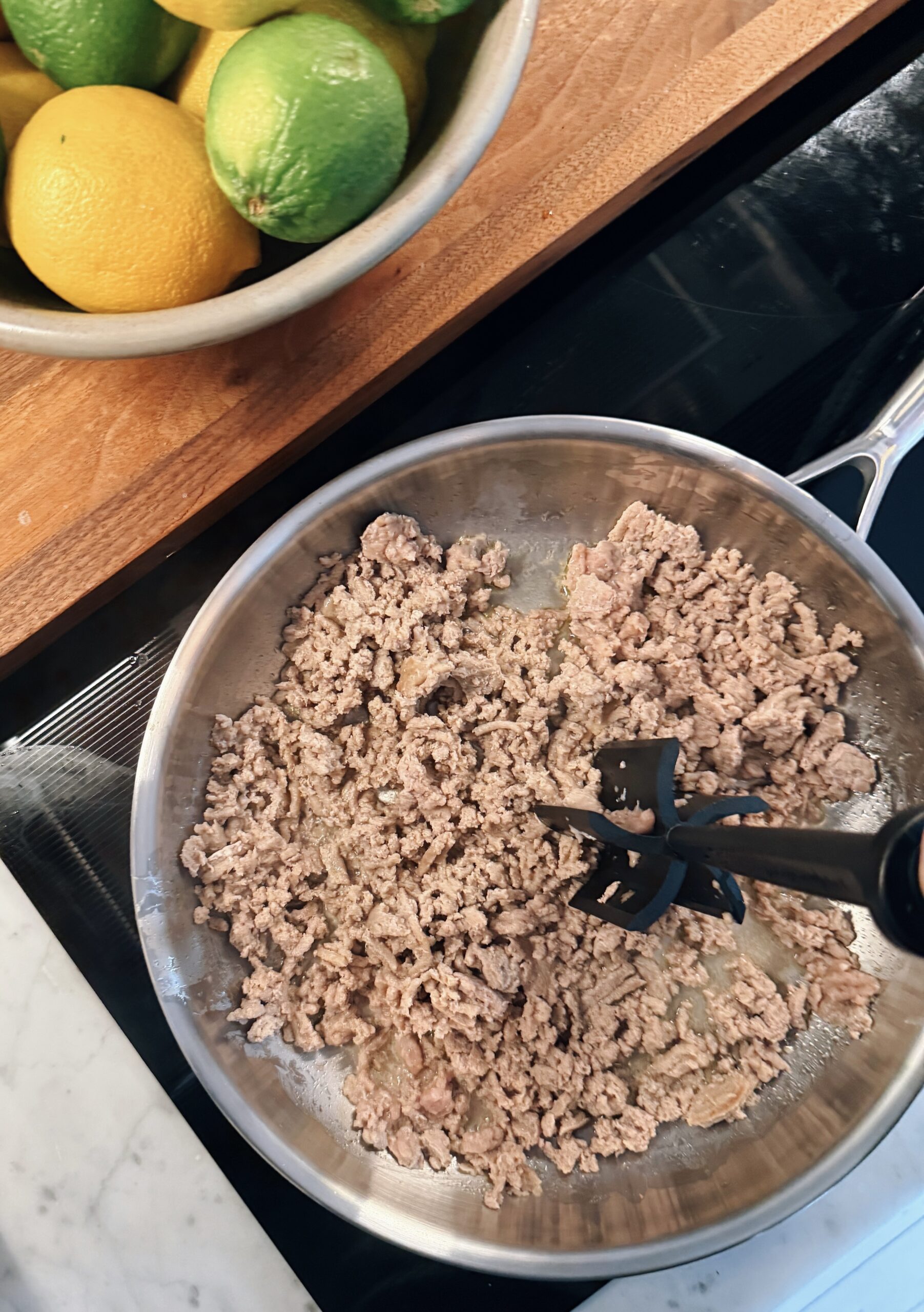 Ground meat is being browned in a stainless steel skillet with a black meat chopper. Next to the pan, there is a bowl filled with lemons and limes on a wooden surface.