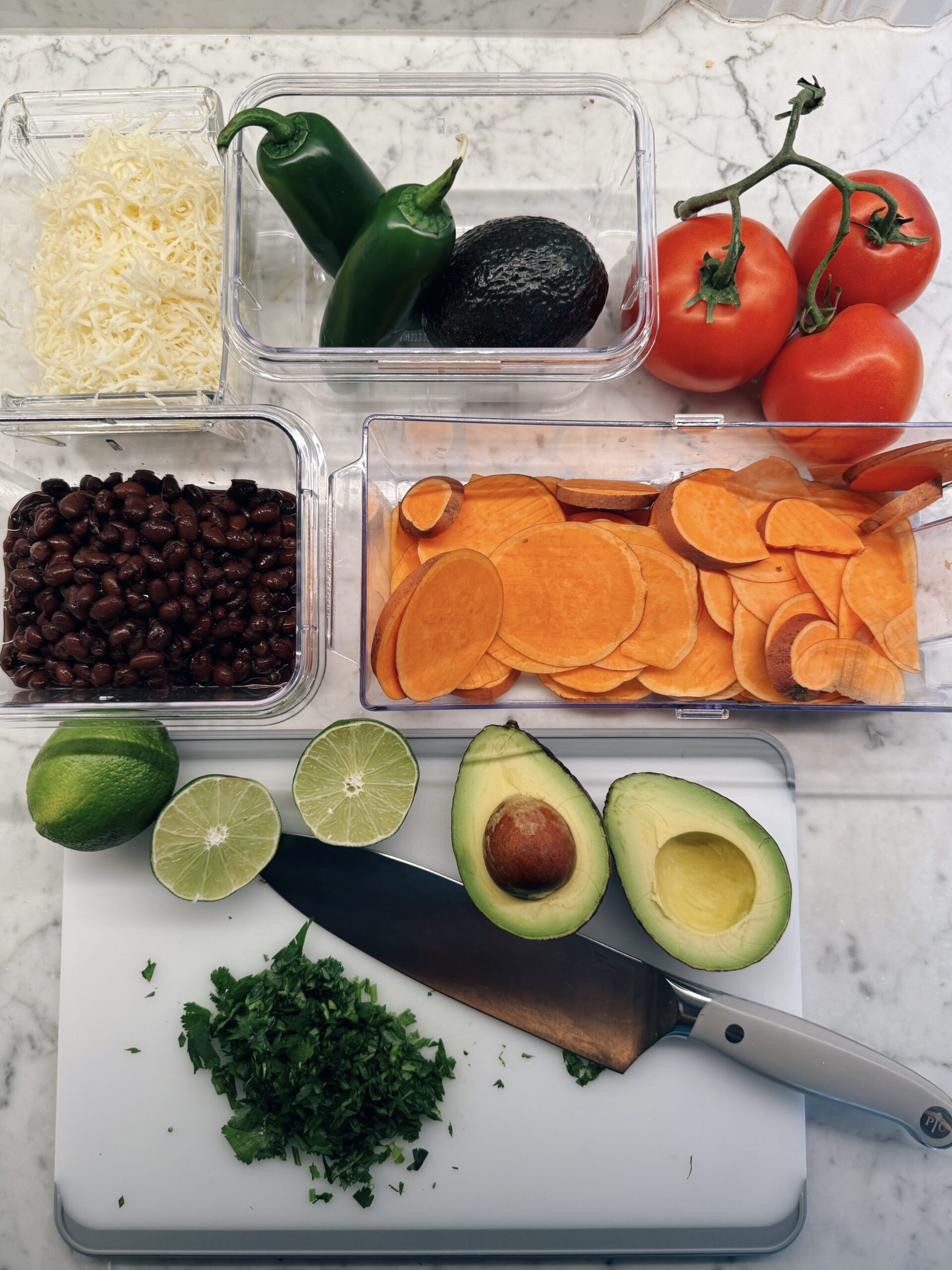 A variety of fresh ingredients are arranged on a white countertop, including sliced sweet potatoes, black beans, cheese, whole tomatoes, jalape&ntilde;os, an avocado, halved limes, and chopped cilantro with a knife on a cutting board.