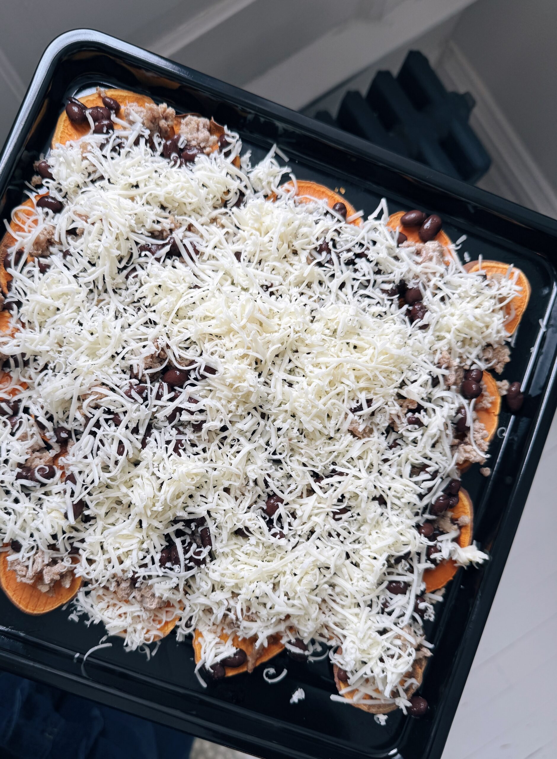 A black tray filled with sliced sweet potatoes topped with black beans, shredded meat, and a generous layer of shredded cheese, ready to be baked.