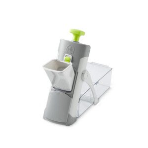 A handheld gray and white kitchen slicer with a green push button, angled chute, and a transparent container for collecting sliced food items.