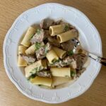 A white plate with rigatoni pasta and pieces of beef in a creamy sauce, garnished with chopped parsley, sits on a wooden table with a fork resting on the plate.