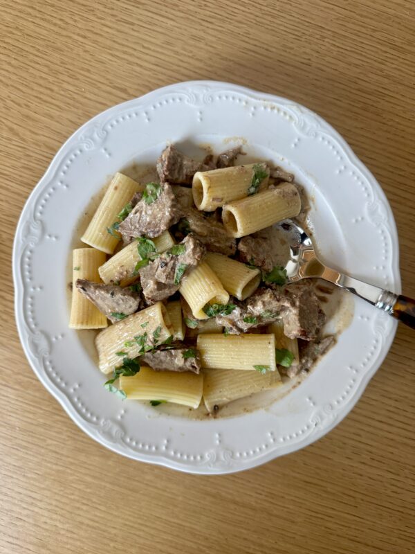 A white plate with rigatoni pasta and pieces of beef in a creamy sauce, garnished with chopped parsley, sits on a wooden table with a fork resting on the plate.
