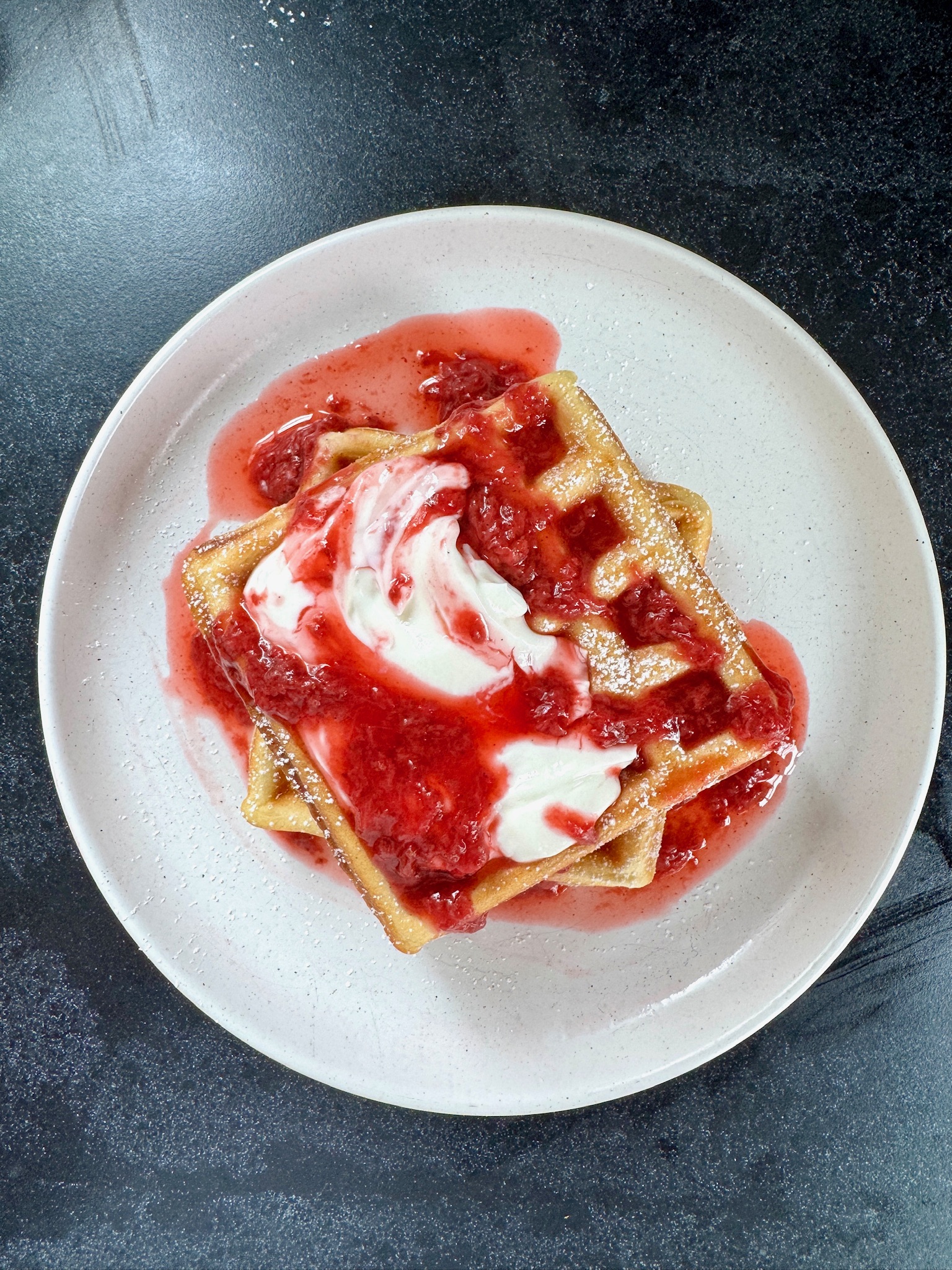 Two golden waffles topped with a swirl of cream and strawberry sauce are served on a white plate, set against a dark background.