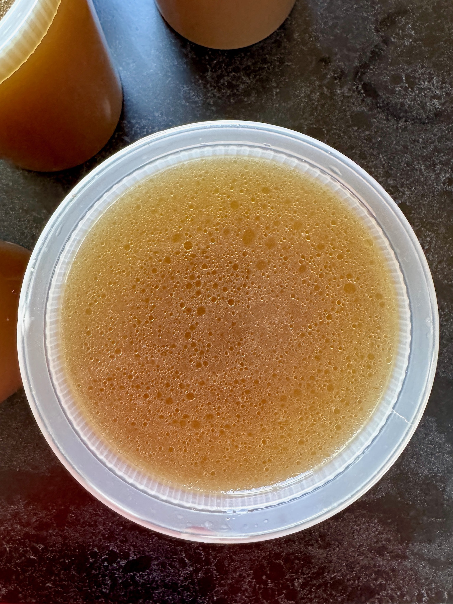 A clear plastic container filled with golden-brown broth sits on a dark countertop, with other similar containers partially visible nearby.