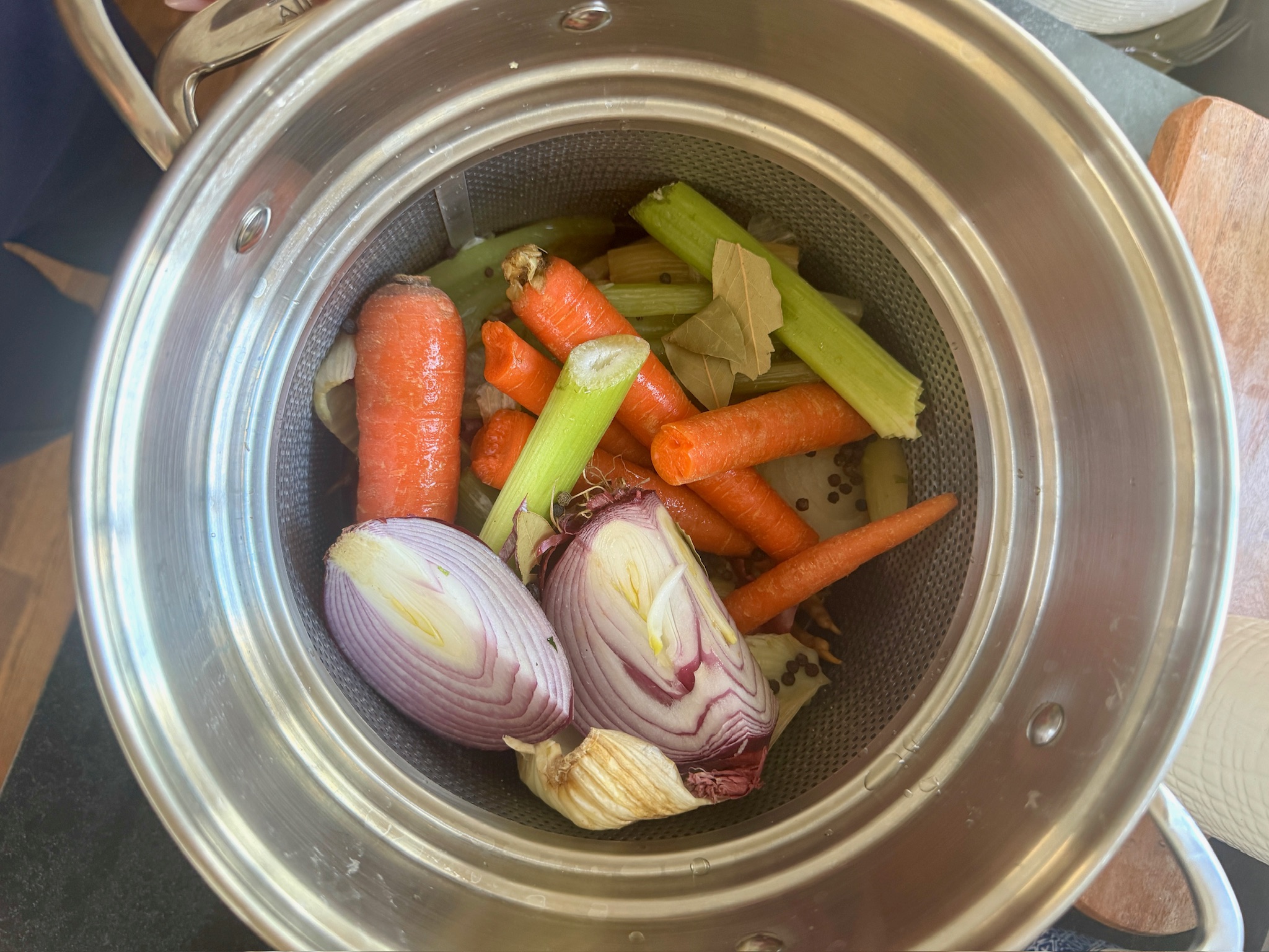 A stainless steel pot filled with vegetables for stock, including halved red onions, whole and chopped carrots, celery sticks, bay leaves, and garlic cloves.