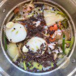 A stainless steel pot filled with water, halved onions, carrots, celery, garlic cloves, peppercorns, and vegetable scraps, prepared for making homemade vegetable broth.