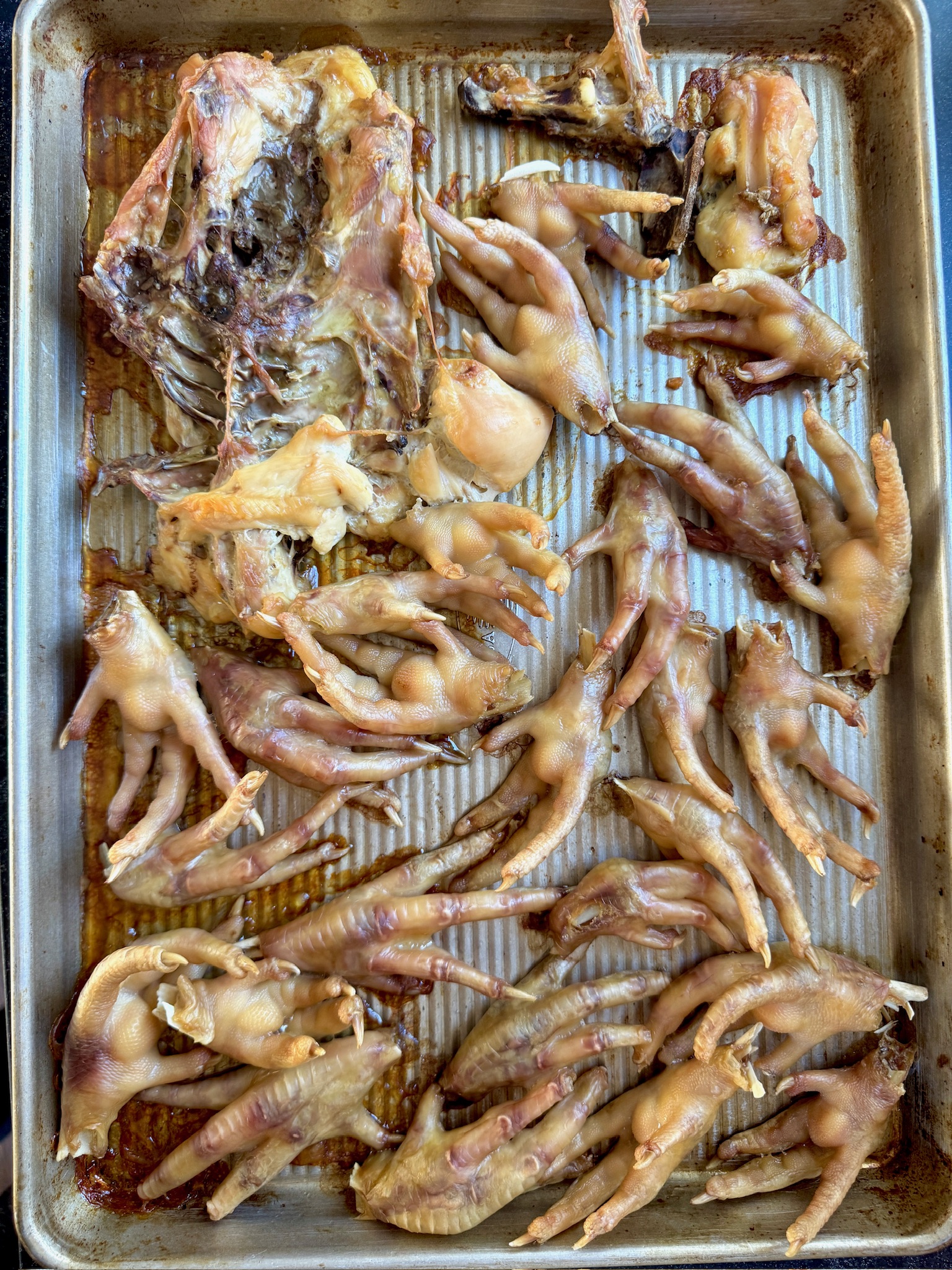 A baking tray filled with cooked chicken feet and a large piece of chicken bone, likely a carcass, arranged on a ridged metal surface. The food appears to be roasted or baked.