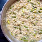 A close-up of creamy risotto in a pan with pieces of green asparagus and yellow zest mixed throughout. The rice looks tender and the dish appears freshly cooked.