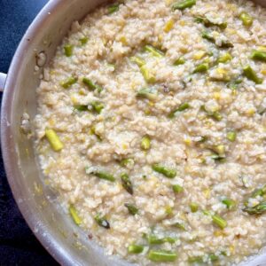 A close-up of creamy risotto in a pan with pieces of green asparagus and yellow zest mixed throughout. The rice looks tender and the dish appears freshly cooked.