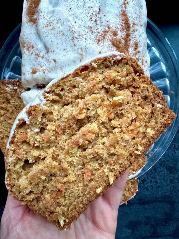 A hand holds a thick slice of moist carrot cake with visible orange shreds, topped with white icing and a dusting of cinnamon, with more cake in the background on a glass plate.