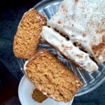 A loaf of carrot cake with white icing and a sprinkle of cinnamon sits on a glass platter. Two thick slices are cut, with one on a white plate below. The cake looks moist and textured.