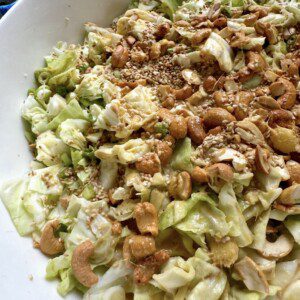 A close-up of a salad in a white bowl, featuring chopped cabbage, cashews, sesame seeds, and a light dressing. The ingredients are mixed and evenly distributed.
