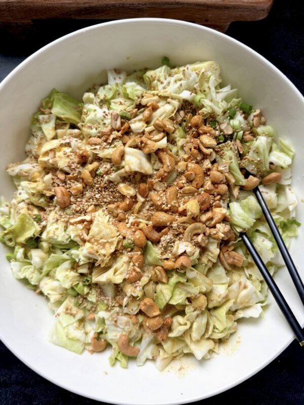 A large white bowl filled with chopped cabbage salad topped with toasted cashews, sesame seeds, and seasoning. A pair of black chopsticks rests on the side of the bowl.