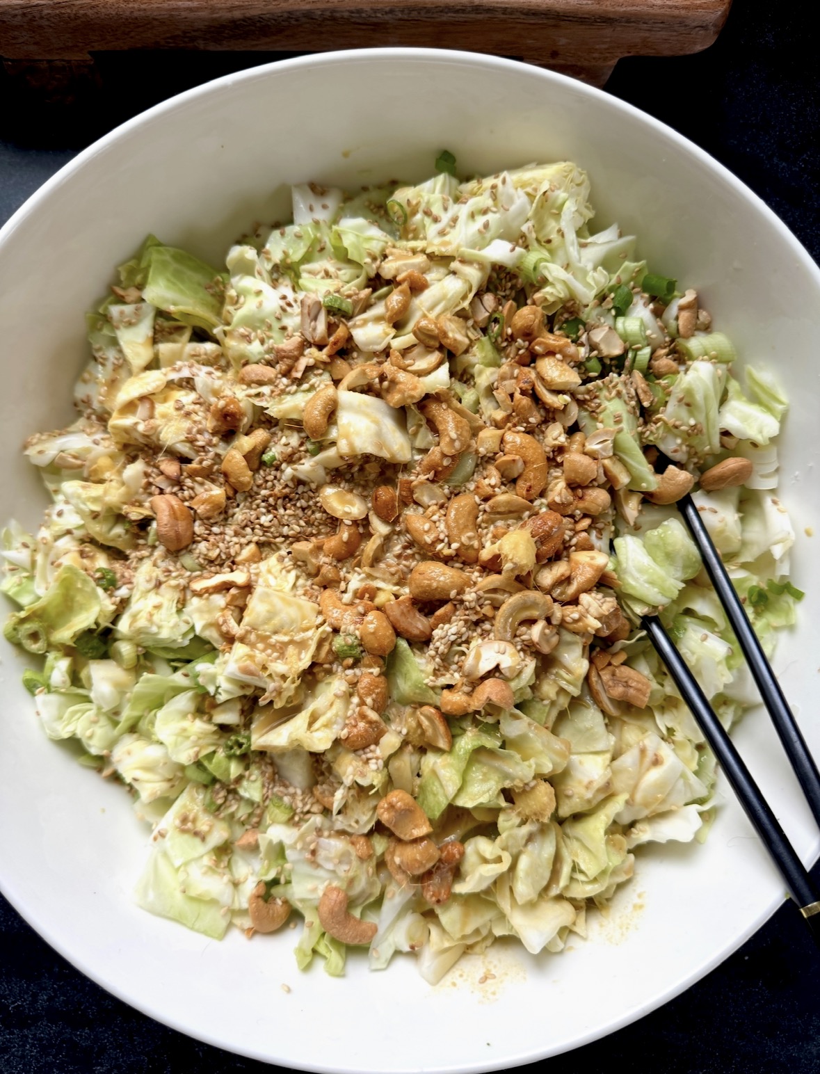 A large white bowl filled with chopped cabbage salad topped with toasted cashews, sesame seeds, and seasoning. A pair of black chopsticks rests on the side of the bowl.