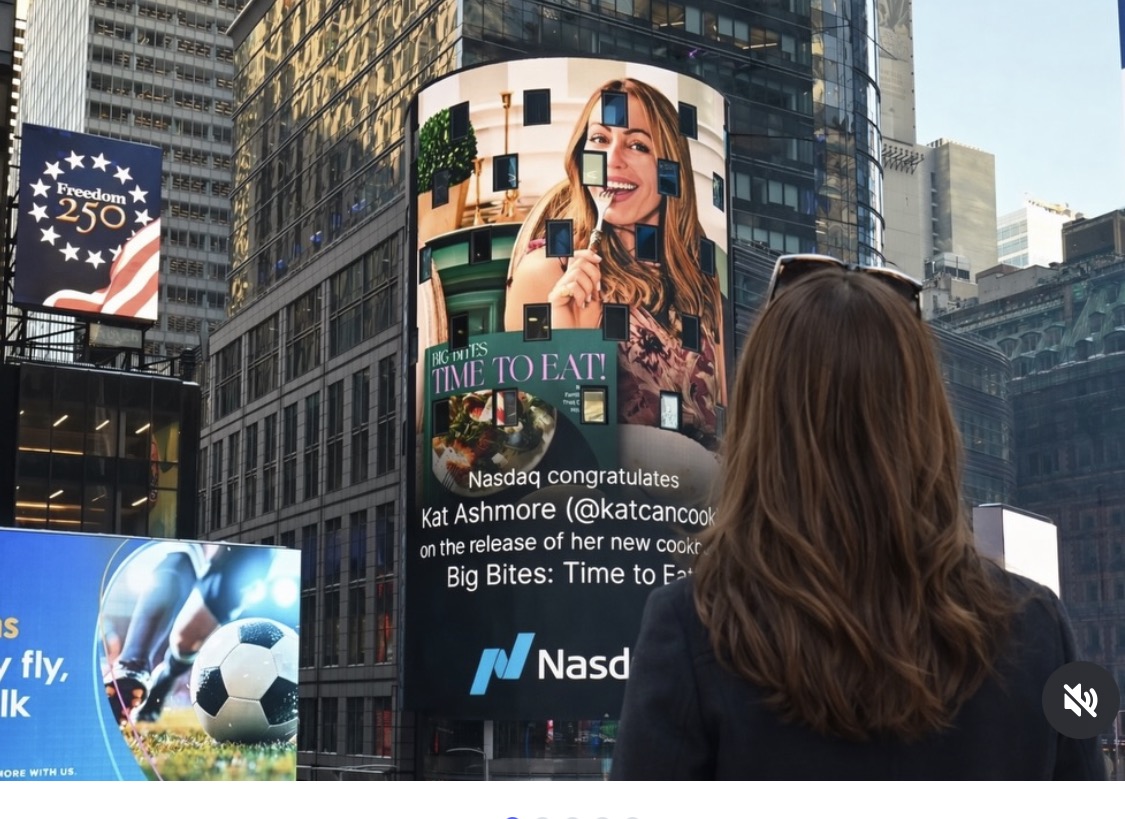 A woman with long brown hair stands in Times Square, looking at a large digital billboard featuring a congratulatory Nasdaq message for Kat Ashmore and the release of her cookbook, with skyscrapers in the background.