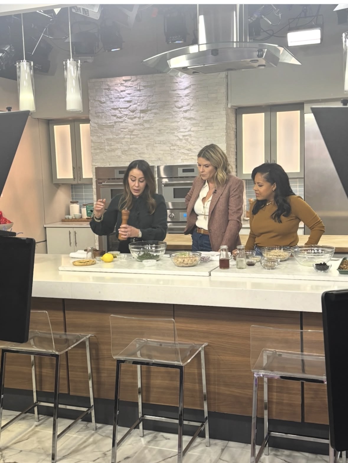 Three women stand in a modern kitchen studio. One is preparing food at the counter while the other two watch. Various ingredients, bowls, and utensils are on the counter. Bright studio lights are visible above.