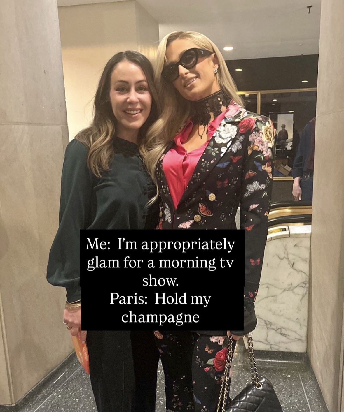 Two women pose together indoors. The woman on the left wears a dark blouse and the woman on the right is dressed glamorously in a floral suit, sunglasses, and a bright shirt. Caption: Me: I&rsquo;m appropriately glam for a morning tv show. Paris: Hold my champagne.
