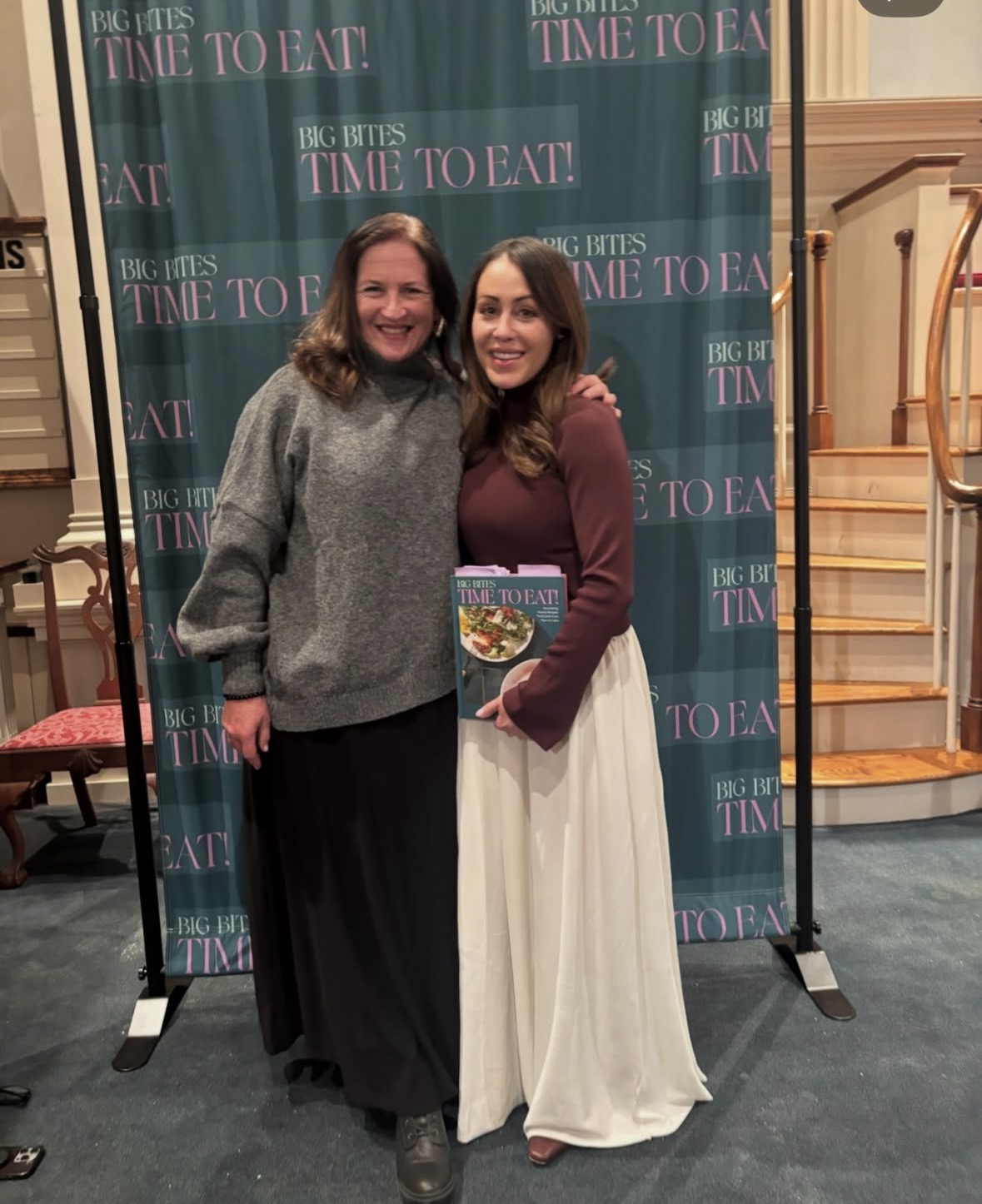 Two women stand smiling in front of a &ldquo;BIG BITES TIME TO EAT!&rdquo; backdrop. One woman holds a cookbook with a dish on the cover. They are indoors near a staircase and wear long skirts and sweaters.