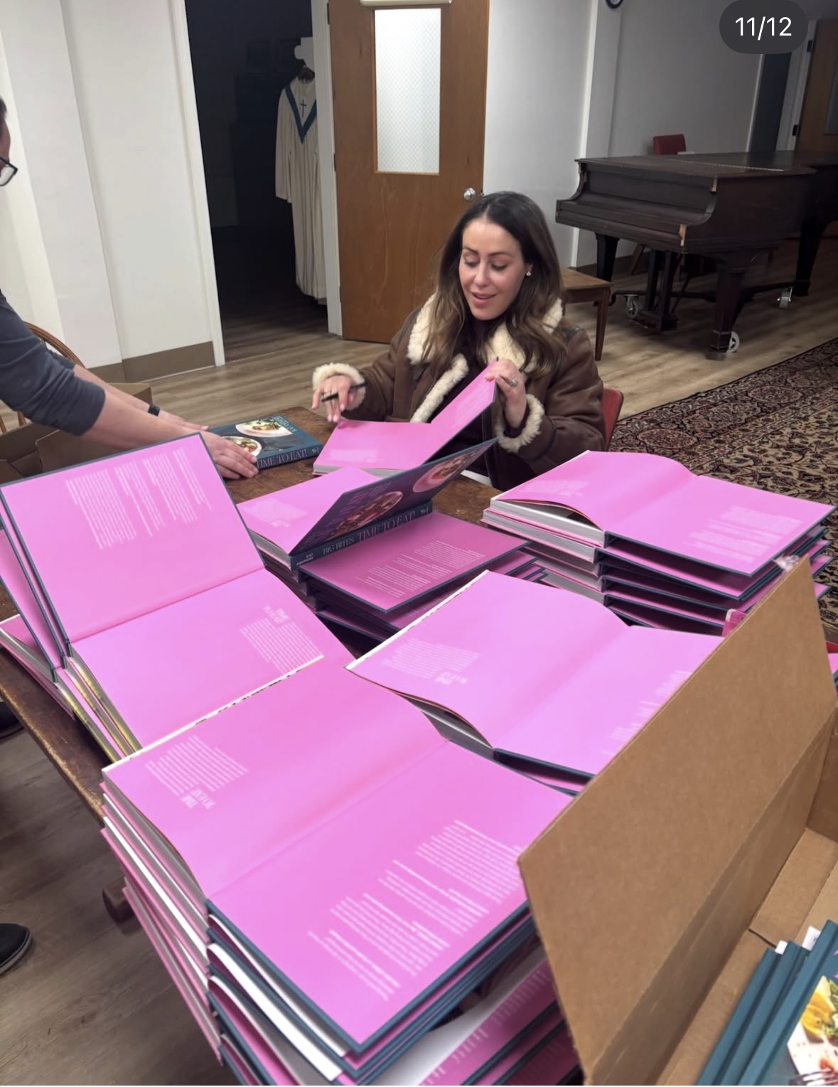 A woman sits at a table stacked with large pink-covered books, flipping through one. Another person&rsquo;s hand is visible, holding a book. A piano and door are in the background in a well-lit room.