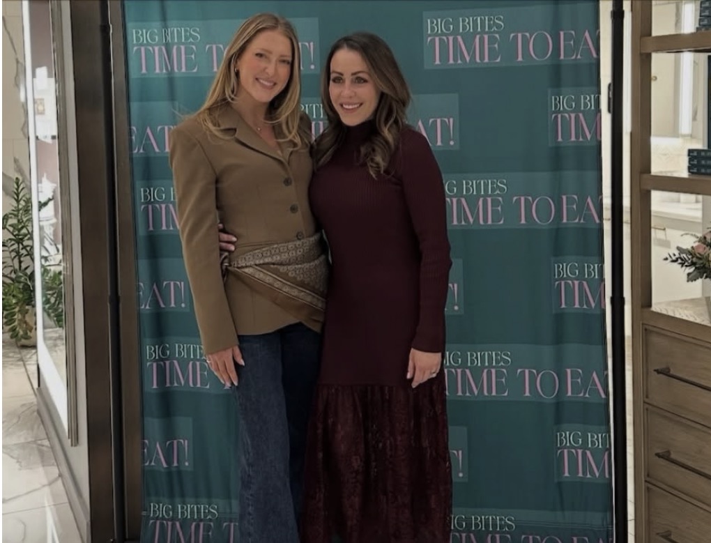Two women pose together and smile in front of a teal photo backdrop with the repeated text, BIG BITES TIME TO EAT! Both are dressed stylishly in casual, elegant outfits.