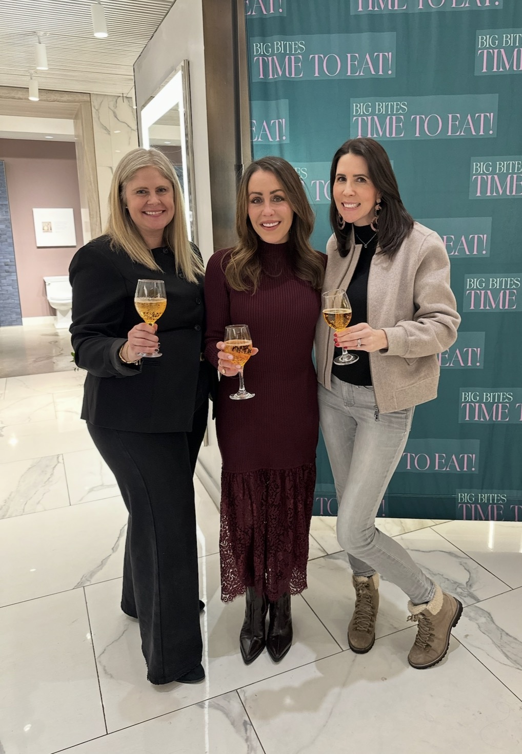 Three women stand together indoors, smiling and holding glasses of sparkling drinks. They pose in front of a green backdrop that reads BIG BITES TIME TO EAT! in a well-lit, modern space with marble floors.