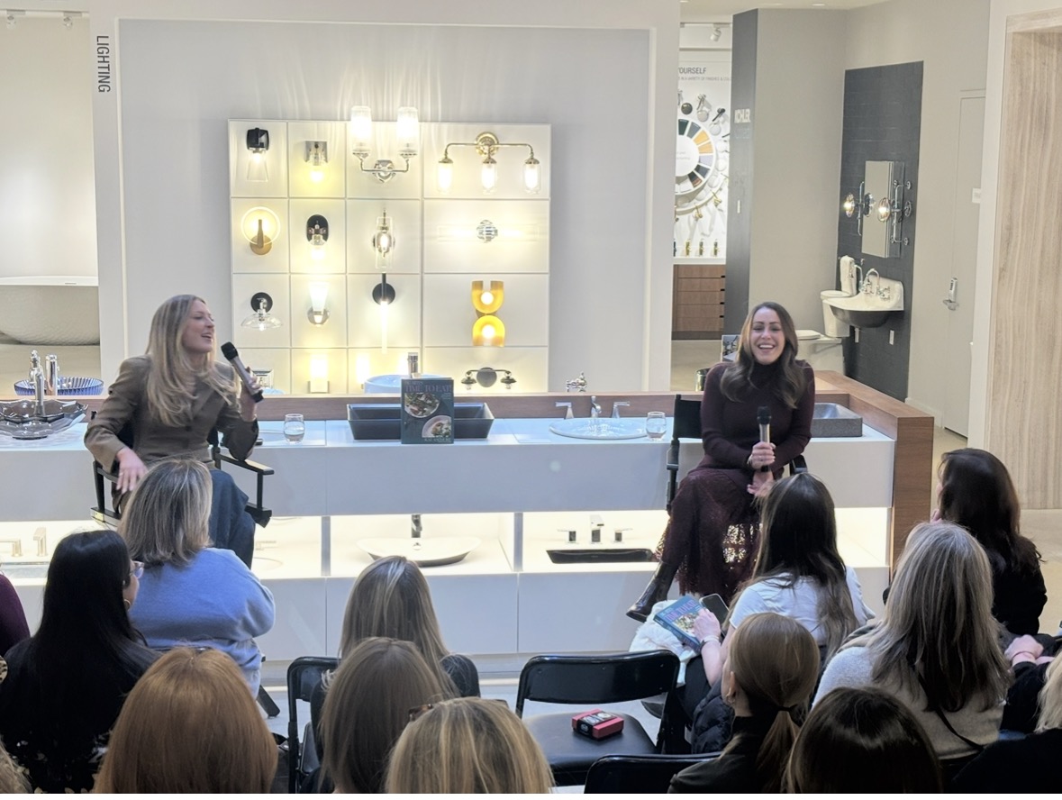 Two women sit facing an audience, smiling and holding microphones. Behind them is a display wall with various light fixtures. The audience is seated and listening attentively in a bright, modern showroom.