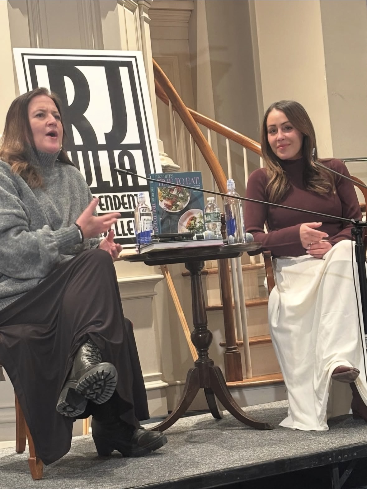 Two women sit and talk on stage beside a small table with books and water bottles, in front of a sign that reads RJ Julia Independent. There is a staircase and part of a banister in the background.