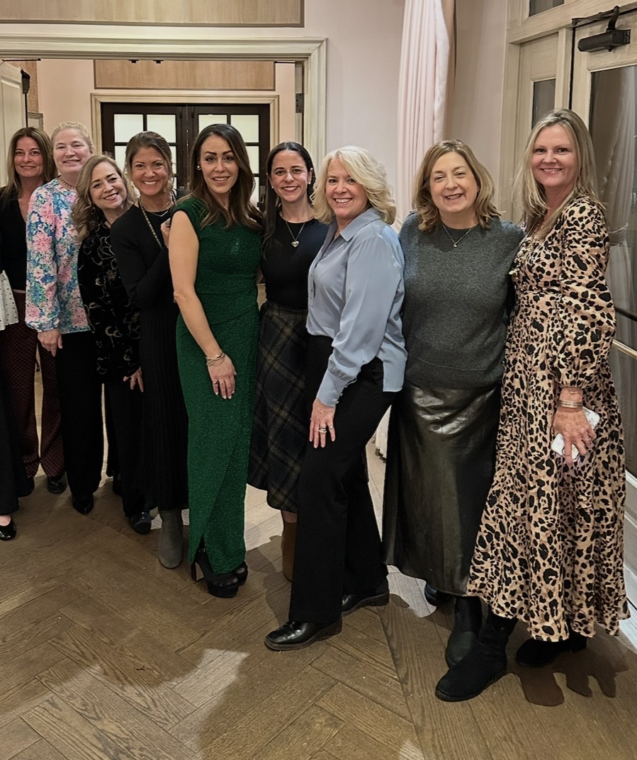 A group of nine smiling women stand together indoors, dressed in a variety of stylish outfits including dresses, skirts, and blouses. They pose closely, with arms around each other, in a warmly lit room with wooden floors.