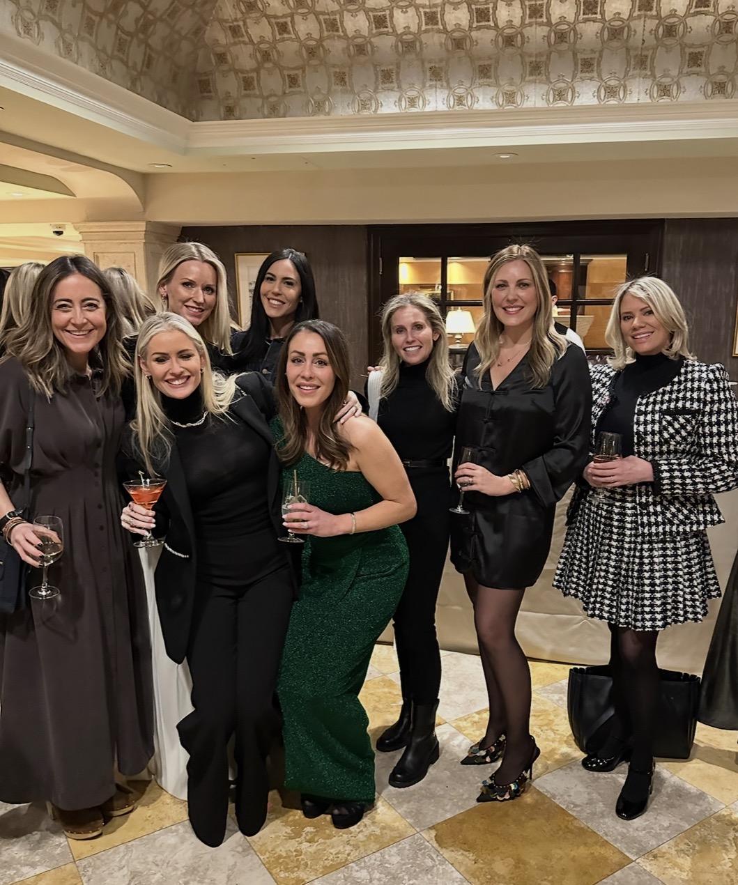 Eight women, dressed in stylish evening attire, stand together indoors, smiling for the camera. Several hold drinks, and the setting features a patterned tile floor and elegant decor.