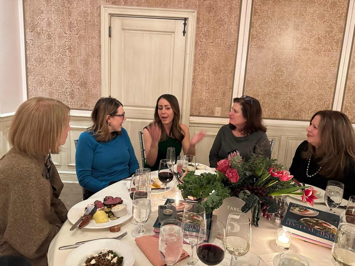 Five women are seated around a dining table, engaged in conversation. There are plates of food, glasses of wine and water, and a floral centerpiece on the table. The setting appears to be a formal or celebratory event.