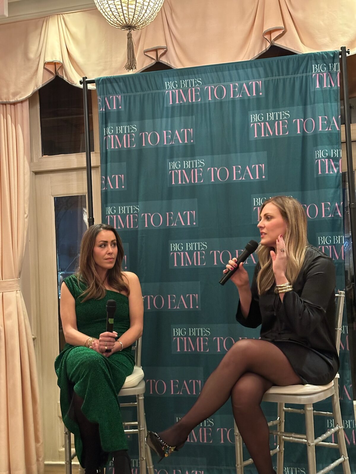 Two women sit on stools, holding microphones, in front of a Big Bites Time To Eat! banner at an indoor event. One woman speaks while the other listens. Both are dressed formally, with a chandelier visible above.