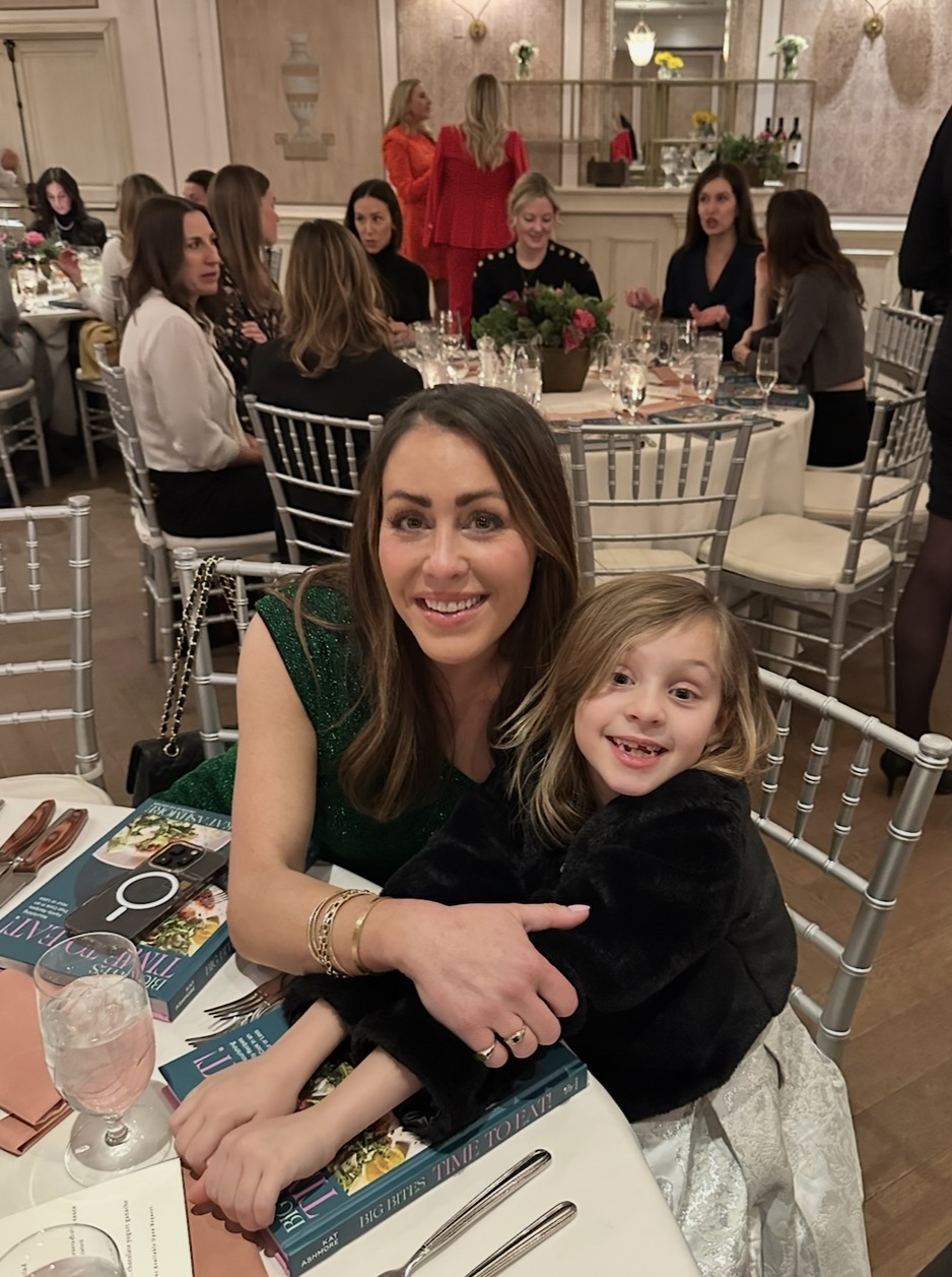 A smiling woman in a green dress hugs a young girl in a black jacket at a dining table set for an event. Other guests sit at tables in the elegant, warmly lit room decorated with flowers.