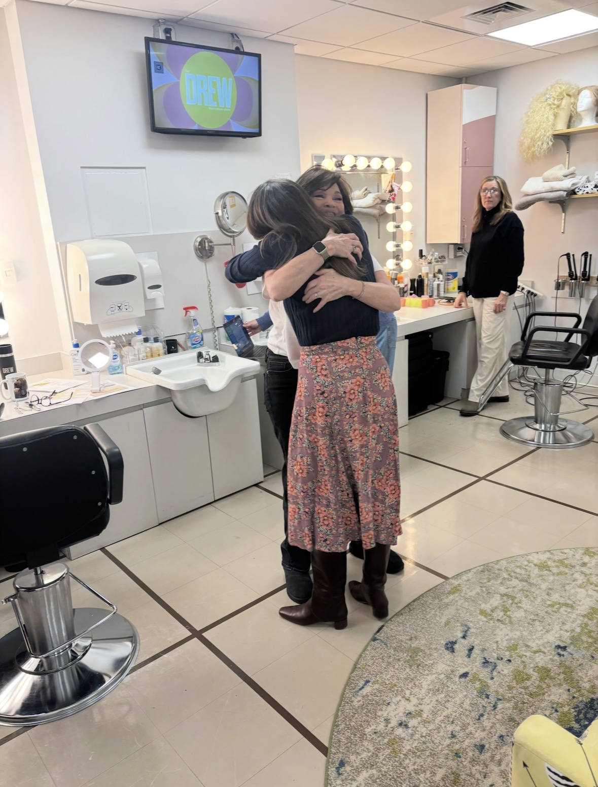Two people hug in a brightly lit dressing room with makeup stations, mirrors, and a TV screen showing DREW in the background. Another person stands nearby, smiling and watching the embrace.