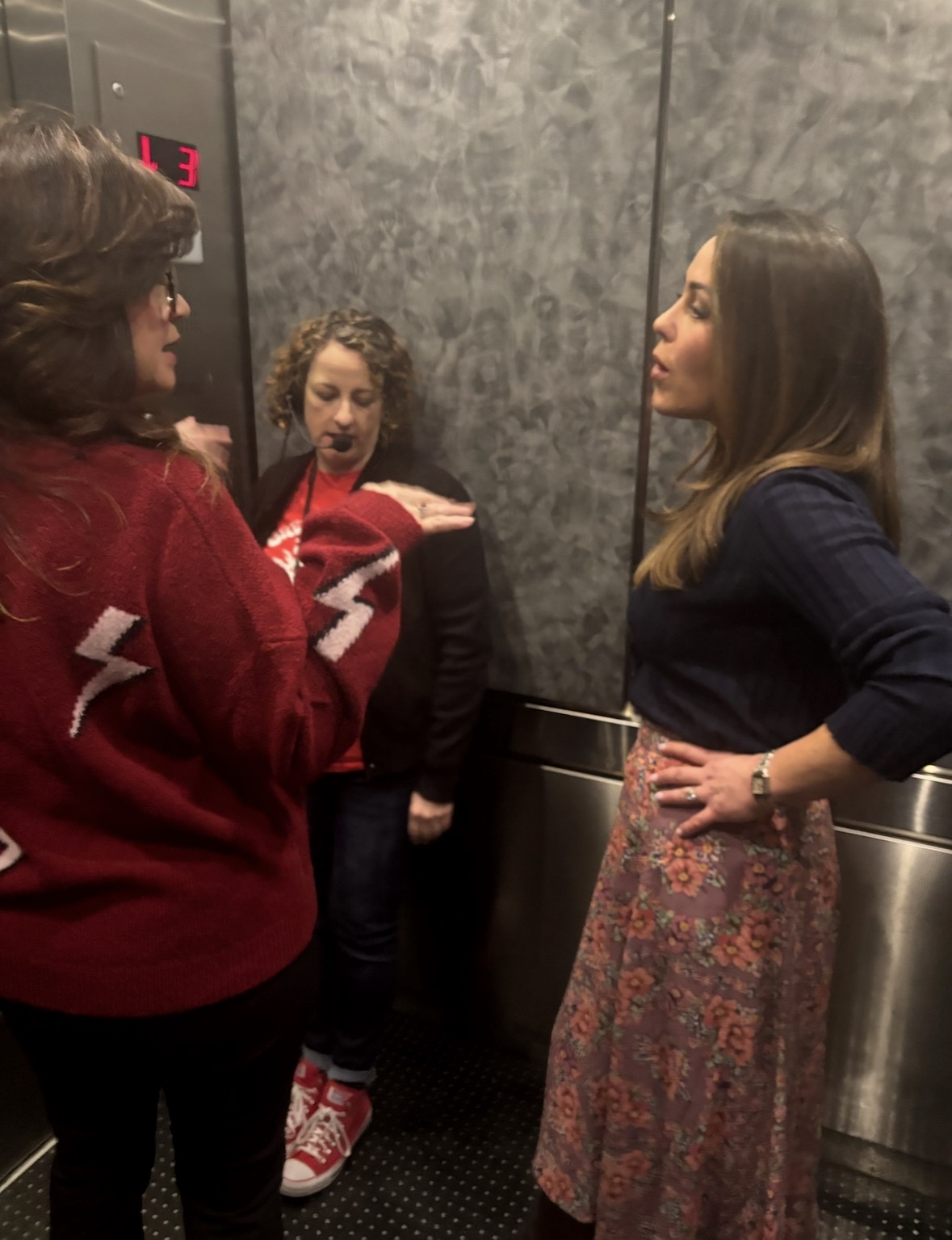 Three women stand in a small elevator having a conversation. One wears a red sweater with white lightning bolts, another wears a navy top and floral skirt, and the third stands in the back wearing black and red sneakers.
