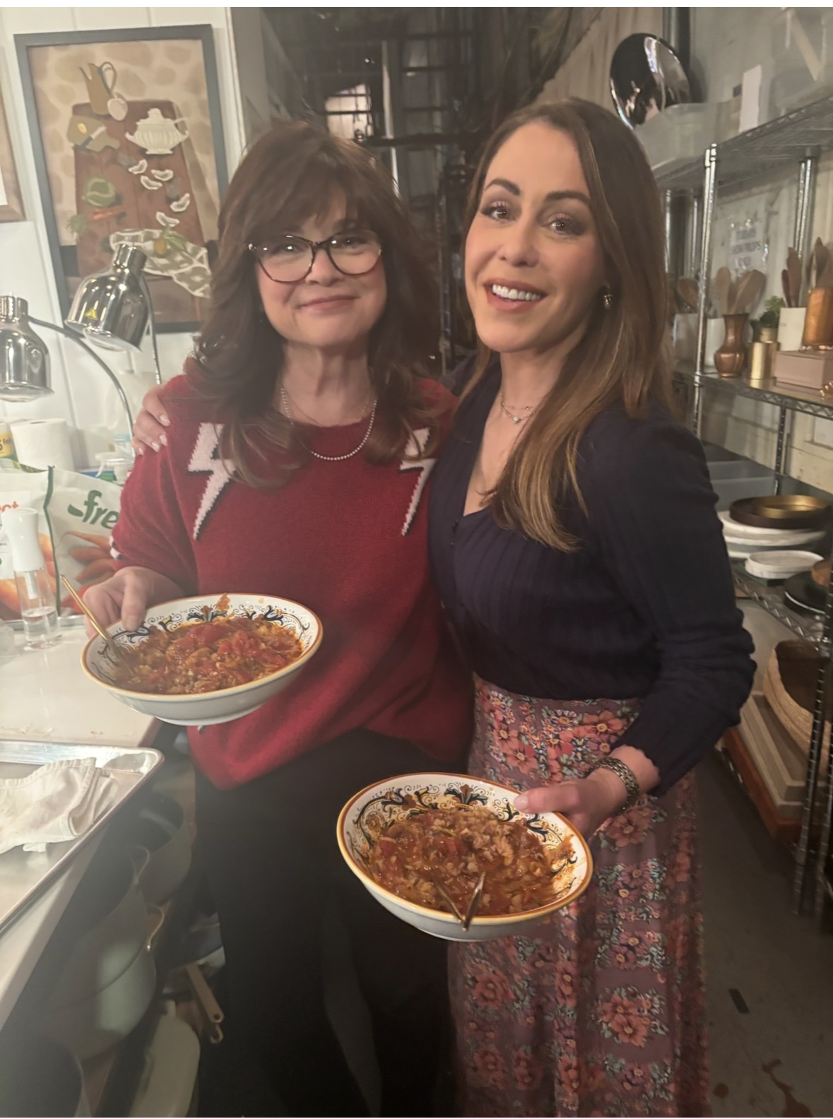 Two women stand side by side in a kitchen, smiling at the camera and holding bowls of pasta with sauce. The background shows shelves with kitchenware and wall art.