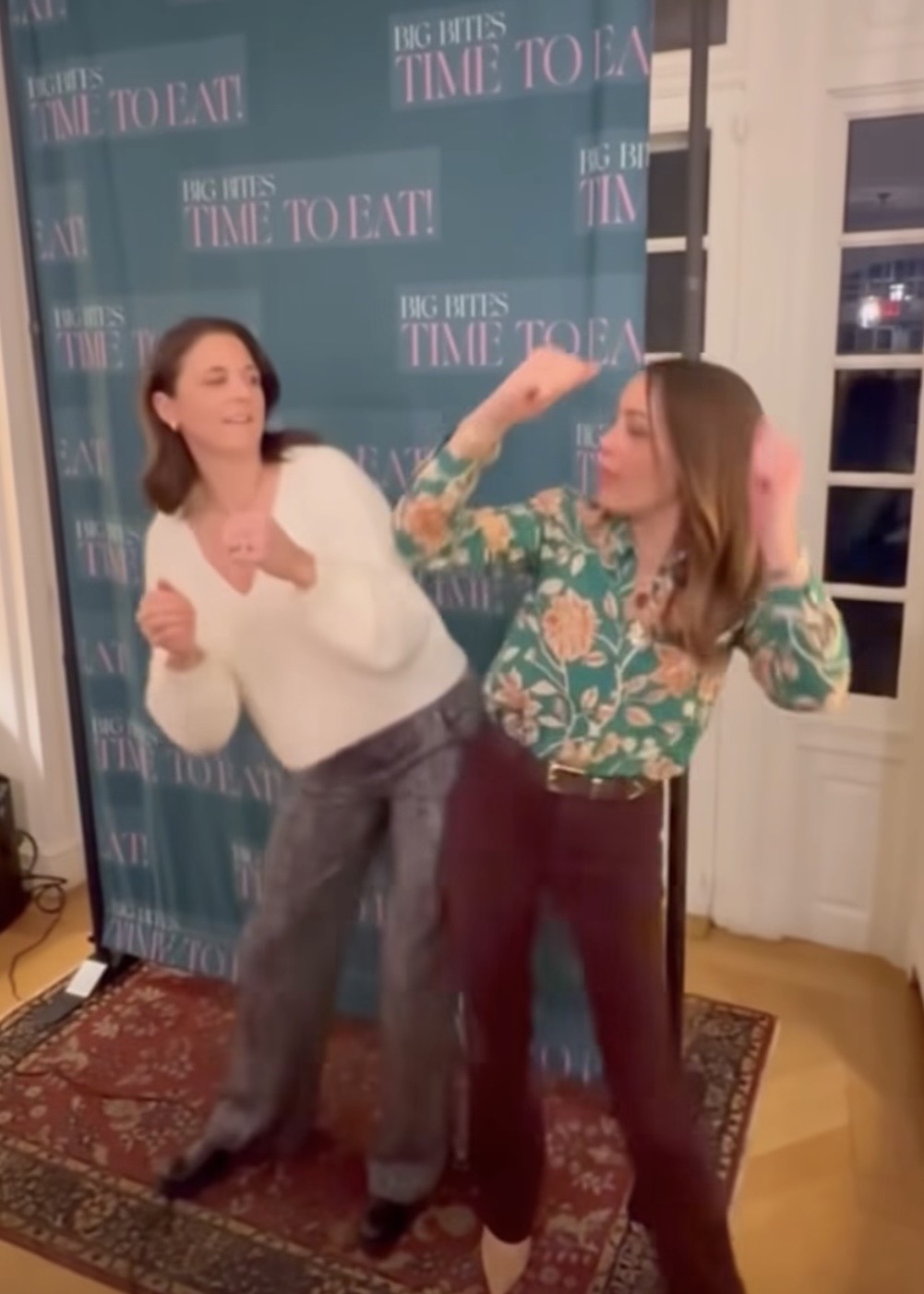 Two women stand in front of a backdrop that says BIG BITES TIME TO EAT! They are playfully posing, with one leaning back and the other raising her arms as if dancing or pretending to box. Both are smiling and having fun.