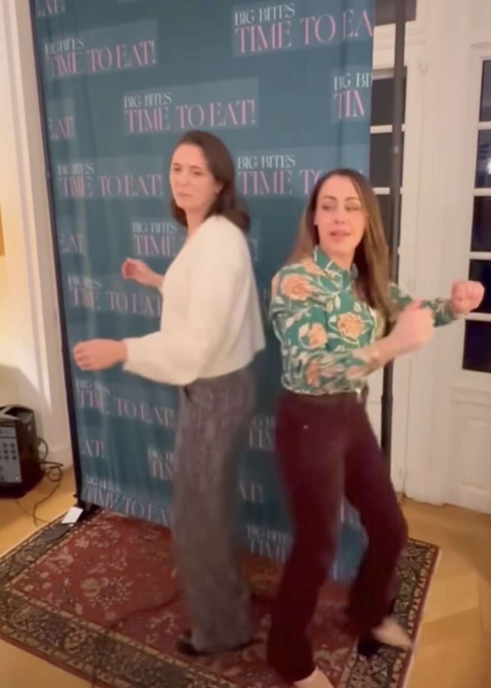Two women dance back-to-back on a patterned rug in front of a blue photo backdrop that reads BIG BITES TIME TO EAT! in a well-lit room.
