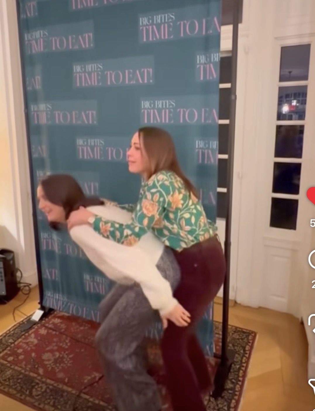 Two women pose playfully in front of a blue backdrop reading BIG BITES TIME TO EAT. One woman leans forward while the other, dressed in a floral shirt, hugs her from behind and appears to be lifting her.