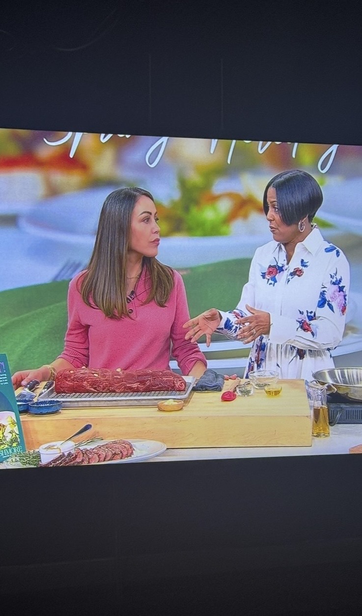 Two women stand behind a kitchen counter preparing food on a cooking show set. One woman wears a pink top and slices meat, while the other, in a white floral blouse, gestures as they talk. Kitchen ingredients are on the counter.