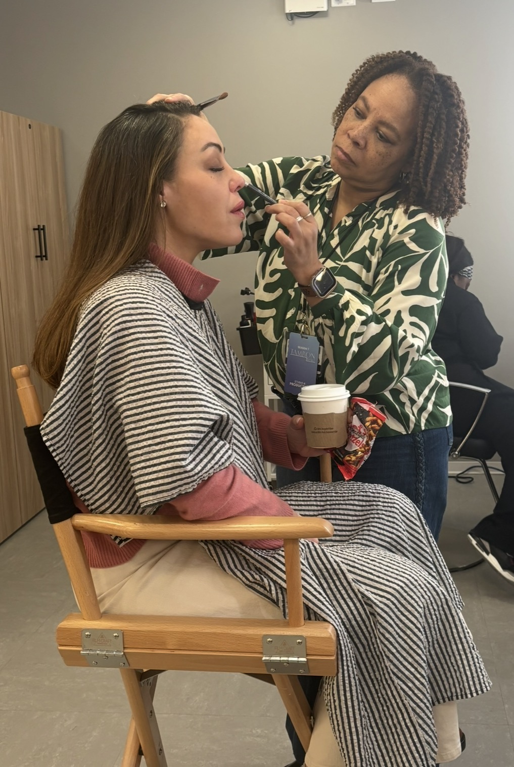 A woman sits in a makeup chair holding a coffee cup while a makeup artist applies makeup to her face. The woman wears a striped cape and a pink sweater; the artist wears a green patterned shirt. Another person is in the background.