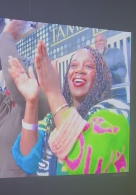 A woman with braided hair smiles and claps, wearing a colorful green, pink, and blue sweater, while standing among a cheering crowd outdoors.
