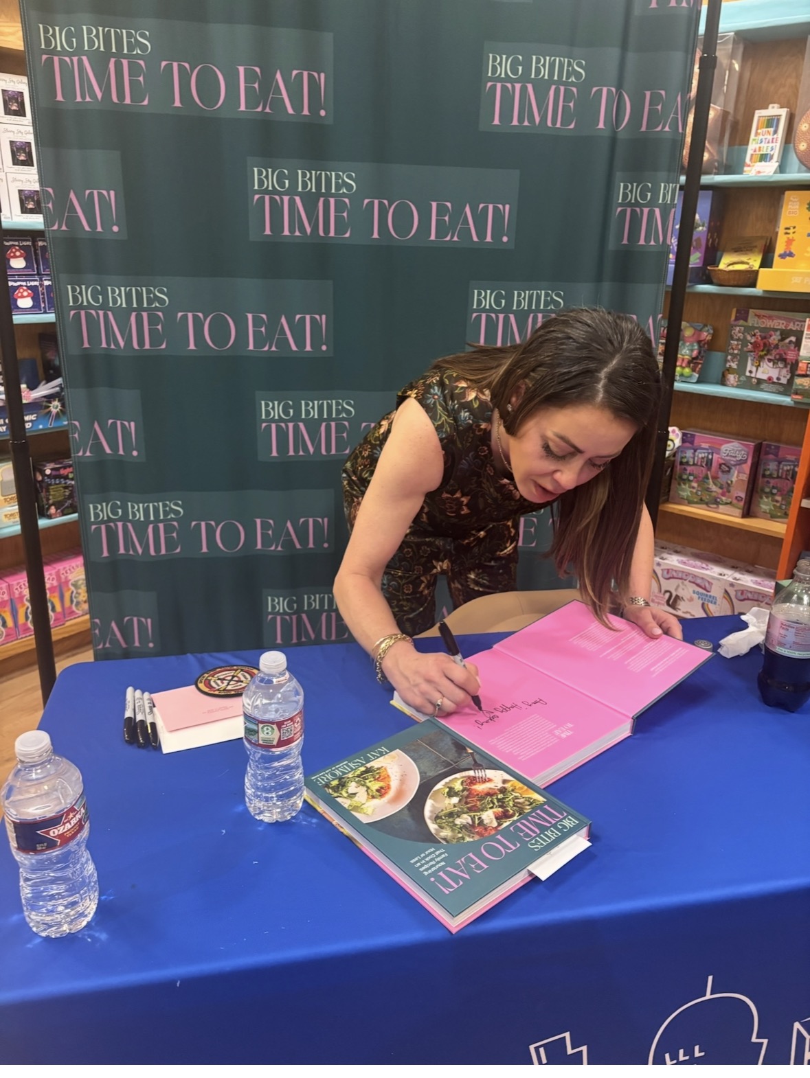 A woman stands at a book signing table, autographing a bright pink book. The backdrop reads BIG BITES TIME TO EAT! and the table displays water bottles, markers, and a cookbook with a photo of a meal on the cover.